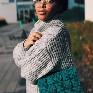 May include: A teal green woven handbag featuring a thick, chunky knit texture and a silver-toned metal chain strap. The bag has a rectangular shape with a distinct grid-like pattern created by the interlaced yarn strands throughout the exterior.