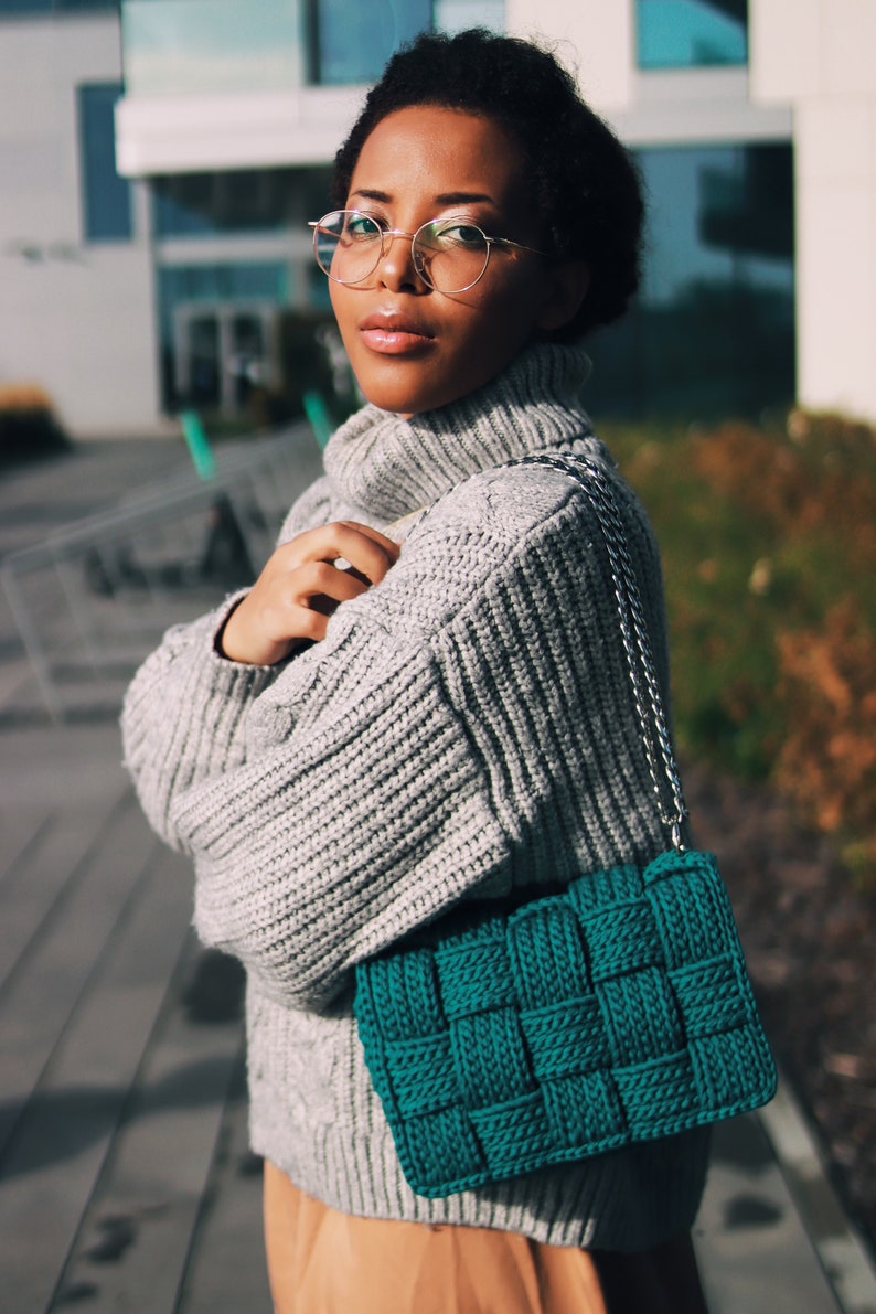 May include: A teal green woven handbag featuring a thick, chunky knit texture and a silver-toned metal chain strap. The bag has a rectangular shape with a distinct grid-like pattern created by the interlaced yarn strands throughout the exterior.