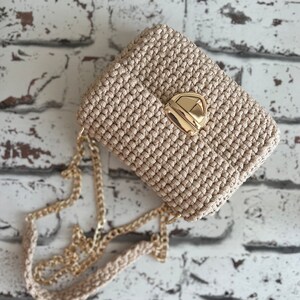 K&ouml;nnte beinhalten: Eine beige geh&auml;kelte Handtasche mit einem strukturierten, geknoteten Muster und einem goldfarbenen Metall-Druckverschluss. Die Tasche verf&uuml;gt &uuml;ber einen passenden beigen H&auml;kel-Schulterriemen mit integrierter goldfarbener Metallkette.