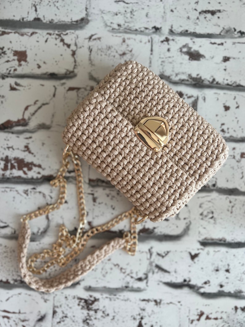 K&ouml;nnte beinhalten: Eine beige geh&auml;kelte Handtasche mit einem strukturierten, geknoteten Muster und einem goldfarbenen Metall-Druckverschluss. Die Tasche verf&uuml;gt &uuml;ber einen passenden beigen H&auml;kel-Schulterriemen mit integrierter goldfarbener Metallkette.