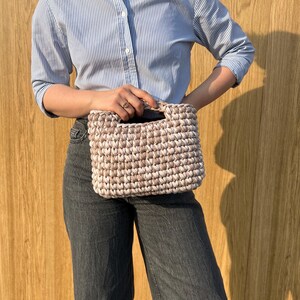 May include: A rectangular handbag crafted from thick, beige and white variegated yarn in a chunky crochet stitch. The bag features a structured, integrated top handle and a dense, textured pattern throughout its exterior surface.