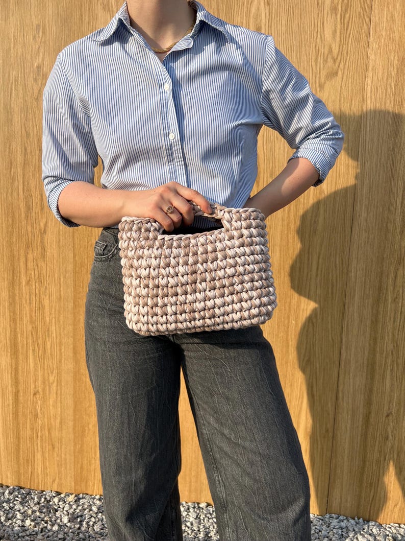 May include: A rectangular handbag crafted from thick, beige and white variegated yarn in a chunky crochet stitch. The bag features a structured, integrated top handle and a dense, textured pattern throughout its exterior surface.