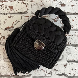 Puede incluir: Un bolso de mano negro de ganchillo con un asa trenzada gruesa, una borla grande a juego en el lateral y un cierre de presi&oacute;n de metal plateado en la solapa frontal. El bolso est&aacute; hecho de hilo de tela grueso con un punto texturizado.