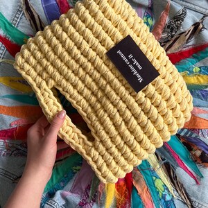 May include: A bright yellow, hand-crocheted handbag made from thick, chunky yarn. The bag features a rectangular shape with a cutout handle and a black rectangular fabric label stitched to the front that reads, Machine cannot make it.