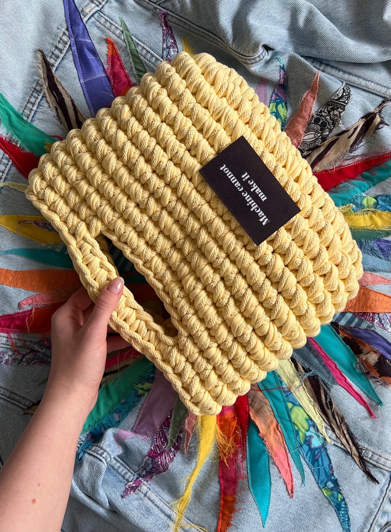 May include: A bright yellow, hand-crocheted handbag made from thick, chunky yarn. The bag features a rectangular shape with a cutout handle and a black rectangular fabric label stitched to the front that reads, Machine cannot make it.