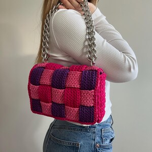 May include: A rectangular handbag featuring a woven pattern of thick, chunky yarn in alternating shades of bright pink and deep purple. The bag is equipped with a heavy, polished silver-tone metal chain strap that connects to the top corners.