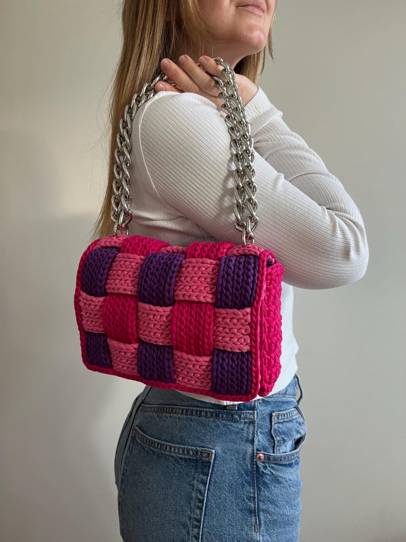 May include: A rectangular handbag featuring a woven pattern of thick, chunky yarn in alternating shades of bright pink and deep purple. The bag is equipped with a heavy, polished silver-tone metal chain strap that connects to the top corners.