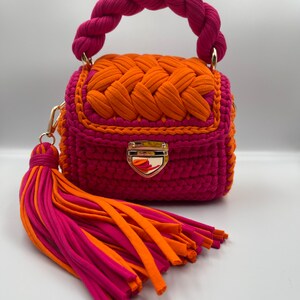 Puede incluir: Un bolso peque&ntilde;o y estructurado hecho de cord&oacute;n de tela rosa vibrante y naranja. Cuenta con un asa superior trenzada, una solapa con un patr&oacute;n naranja tejido, un cierre de metal dorado y una borla grande de m&uacute;ltiples hilos en rosa y naranja.