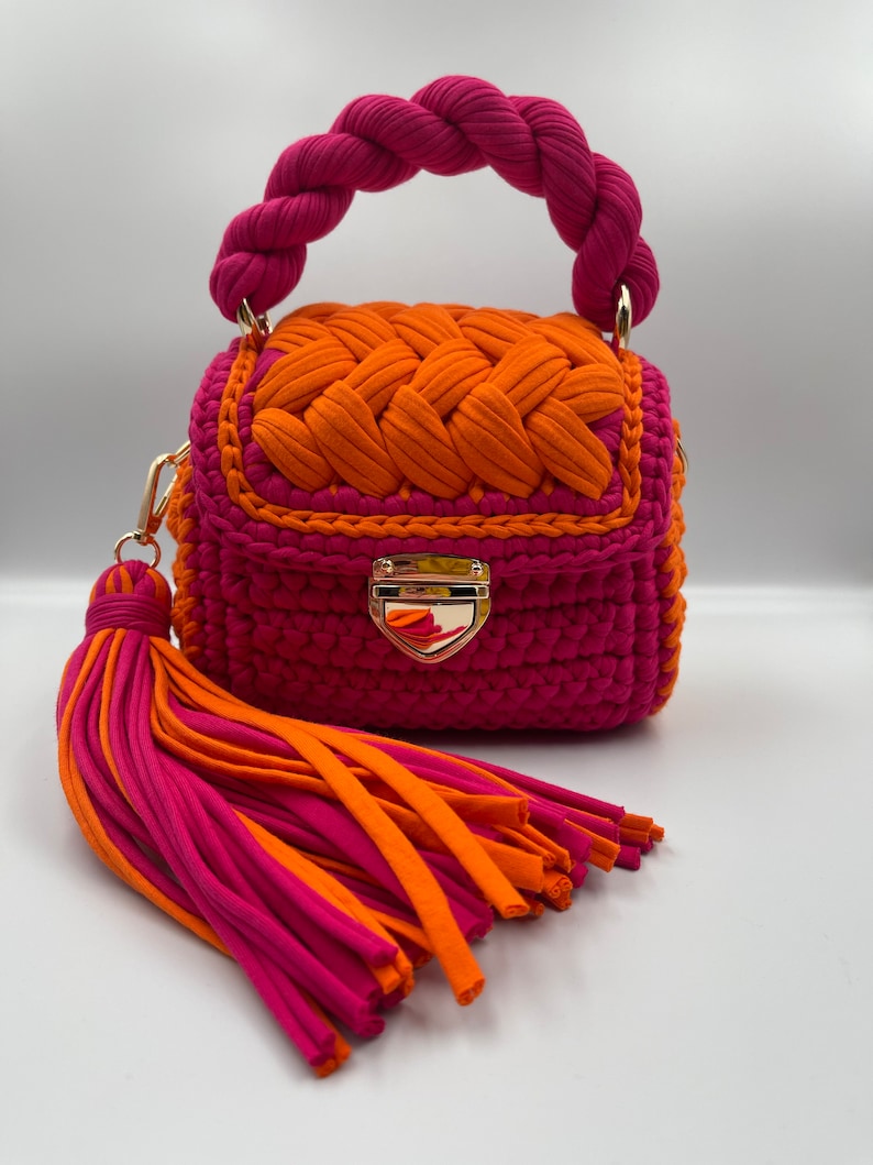 Puede incluir: Un bolso peque&ntilde;o y estructurado hecho de cord&oacute;n de tela rosa vibrante y naranja. Cuenta con un asa superior trenzada, una solapa con un patr&oacute;n naranja tejido, un cierre de metal dorado y una borla grande de m&uacute;ltiples hilos en rosa y naranja.