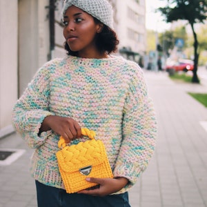 Puede incluir: Una diadema de punto gris claro con un detalle frontal trenzado, un su&eacute;ter de punto grueso con una mezcla de hilos pastel multicolor en rosa, amarillo y azul, y un bolso de ganchillo amarillo brillante con asa trenzada y cierre plateado.