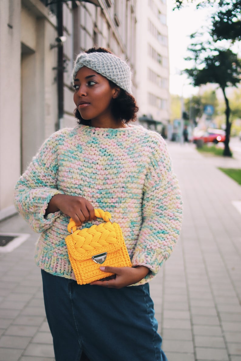 Puede incluir: Una diadema de punto gris claro con un detalle frontal trenzado, un su&eacute;ter de punto grueso con una mezcla de hilos pastel multicolor en rosa, amarillo y azul, y un bolso de ganchillo amarillo brillante con asa trenzada y cierre plateado.