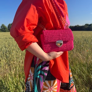 K&ouml;nnte beinhalten: Eine leuchtend pinke H&auml;keltasche mit rechteckiger Klappe, einem goldfarbenen Metalldruckverschluss und einem passenden pinken H&auml;kel-Schulterriemen mit goldfarbenen Kettengliedern, getragen &uuml;ber einem orangefarbenen Hemd und einer gemusterten Hose.