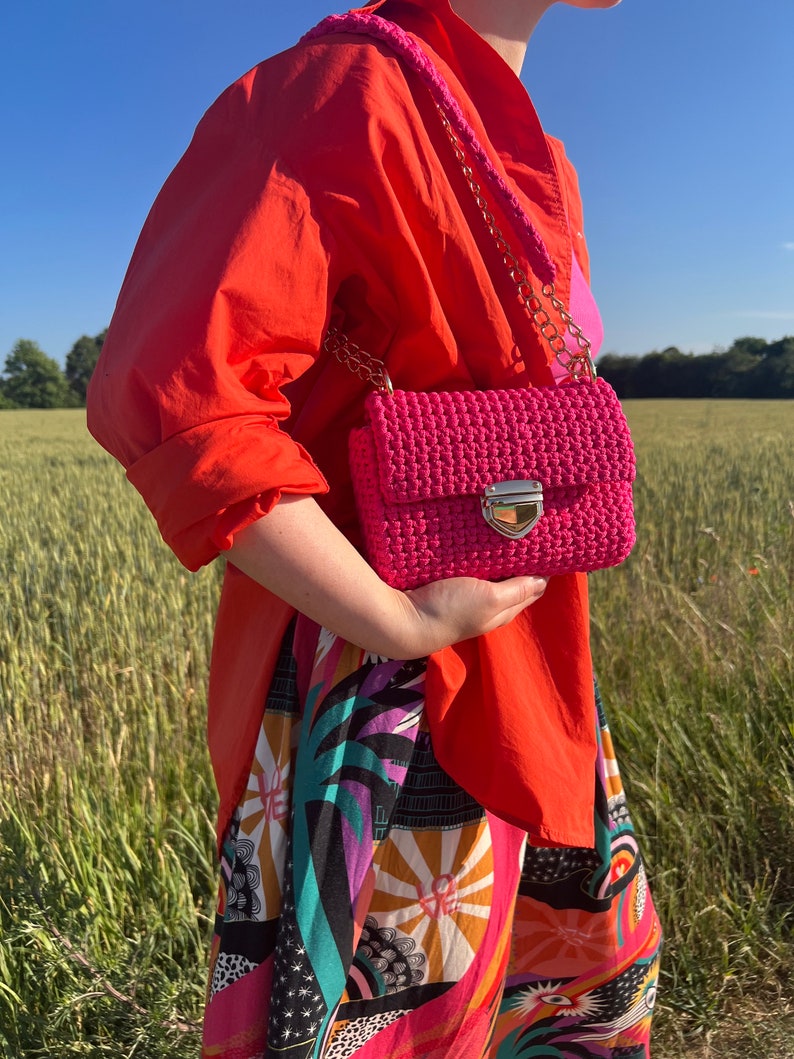 K&ouml;nnte beinhalten: Eine leuchtend pinke H&auml;keltasche mit rechteckiger Klappe, einem goldfarbenen Metalldruckverschluss und einem passenden pinken H&auml;kel-Schulterriemen mit goldfarbenen Kettengliedern, getragen &uuml;ber einem orangefarbenen Hemd und einer gemusterten Hose.