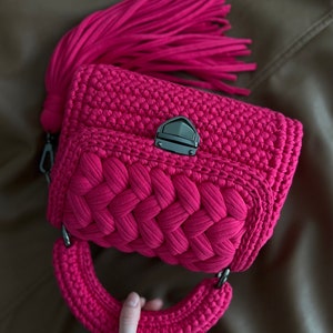 Op de afbeelding: Een levendige fuchsia gehaakte handtas met een dik gevlochten voorpaneel, een stevig rond handvat en een grote bijpassende kwast aan de zijkant. De tas is beveiligd met een donkere metalen druksluiting op de centrale klep.