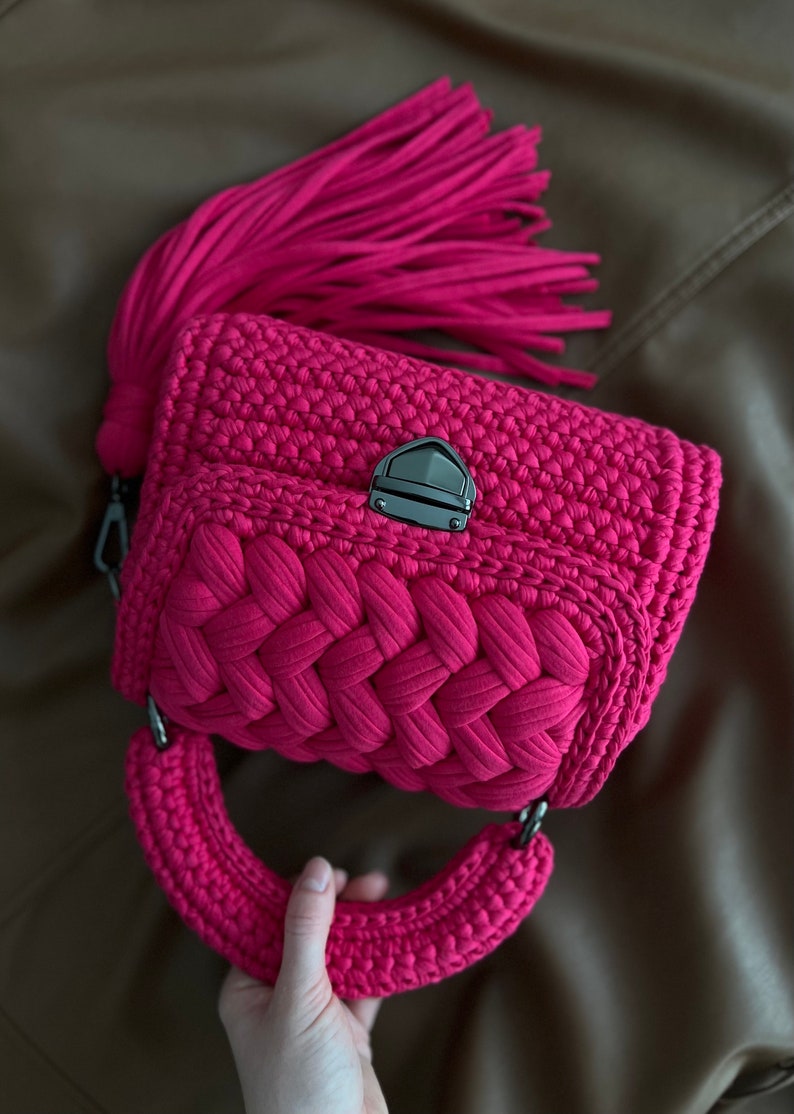 Op de afbeelding: Een levendige fuchsia gehaakte handtas met een dik gevlochten voorpaneel, een stevig rond handvat en een grote bijpassende kwast aan de zijkant. De tas is beveiligd met een donkere metalen druksluiting op de centrale klep.