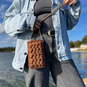 May include: A rectangular brown crochet crossbody bag featuring a thick, braided yarn texture. The bag is suspended by a gold-toned metal chain strap, showcasing a dense, woven pattern that creates a raised, tactile surface across the front.