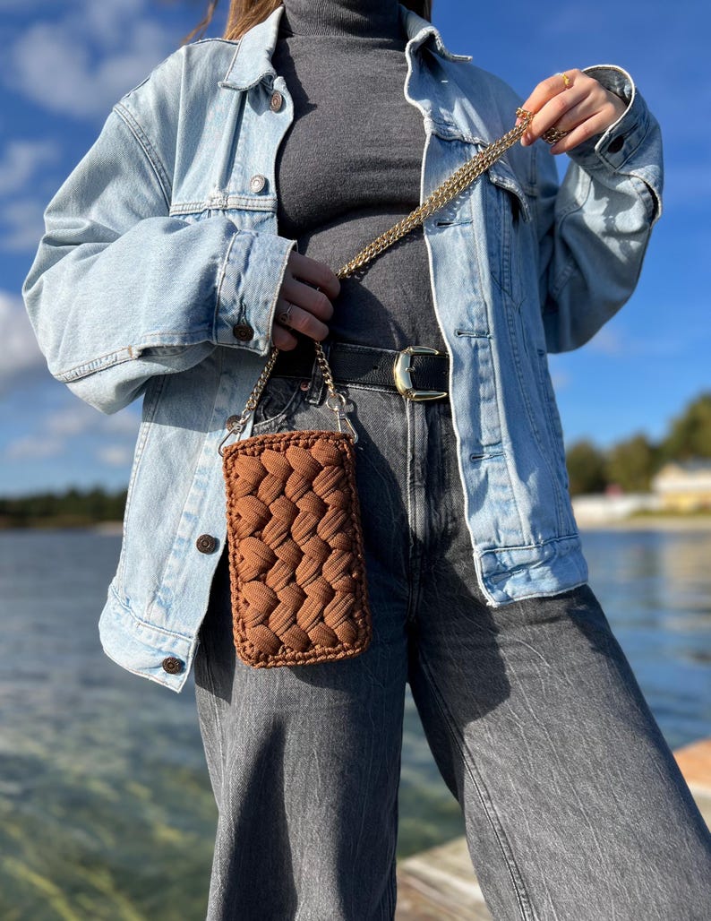 May include: A rectangular brown crochet crossbody bag featuring a thick, braided yarn texture. The bag is suspended by a gold-toned metal chain strap, showcasing a dense, woven pattern that creates a raised, tactile surface across the front.