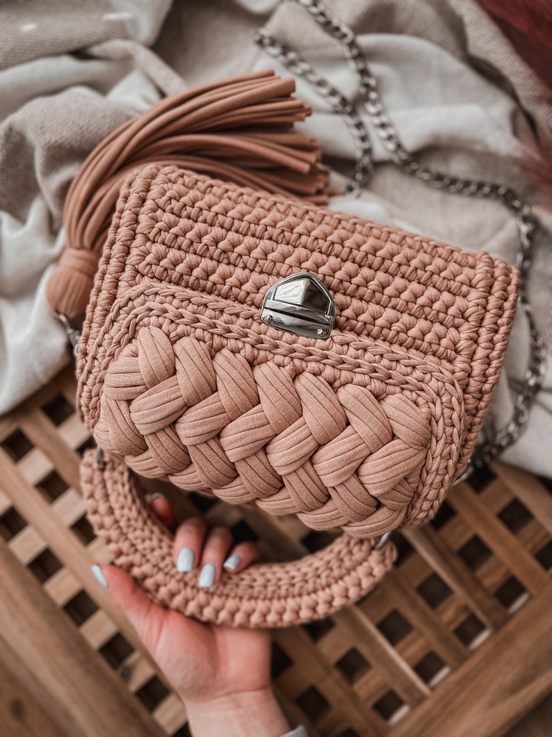 Op de afbeelding: Een oudroze gehaakte handtas met een gevlochten voorpaneel, een zilverkleurige draaisluiting, een bijpassend dik gehaakt handvat en een lange zilveren schouderketting. Aan de zijkant is een grote bijpassende kwast bevestigd.