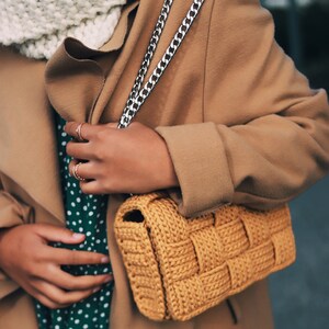 May include: A mustard yellow woven crochet handbag with a silver chain strap, worn with a tan coat, a white chunky knit scarf, and a dark green dress featuring a white polka dot pattern. The bag has a thick, textured, basket-weave design.