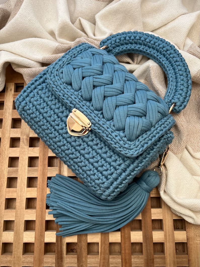 Op de afbeelding: Een stoffig blauwe gehaakte handtas met een gevlochten klepontwerp, een dikke ronde handgreep en een goudkleurige metalen druksluiting. De tas is voorzien van een lange, bijpassende blauwe stoffen kwast die aan de zijkant is bevestigd.