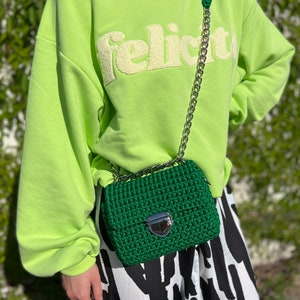 K&ouml;nnte beinhalten: Eine dunkelgr&uuml;ne geh&auml;kelte Handtasche mit silberfarbenem Metallkettenriemen und silbernem Druckknopfverschluss. Die Tasche wird &uuml;ber einem lindgr&uuml;nen Sweatshirt mit dem Wort "felicita" in Reliefschrift und einem schwarz-wei&szlig; gemusterten Rock getragen.