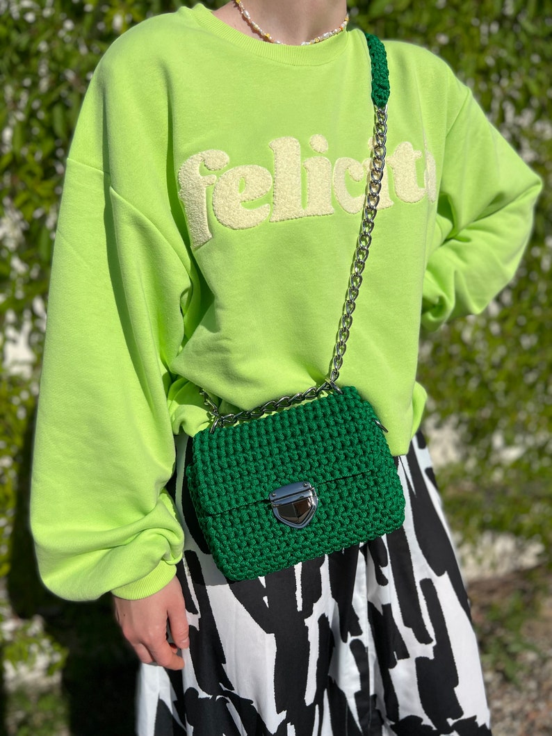 K&ouml;nnte beinhalten: Eine dunkelgr&uuml;ne geh&auml;kelte Handtasche mit silberfarbenem Metallkettenriemen und silbernem Druckknopfverschluss. Die Tasche wird &uuml;ber einem lindgr&uuml;nen Sweatshirt mit dem Wort "felicita" in Reliefschrift und einem schwarz-wei&szlig; gemusterten Rock getragen.