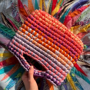 May include: A hand-crocheted bag made from thick fabric yarn featuring a vibrant gradient pattern that transitions from deep purple and lavender at the base to bright pink, orange, and coral tones at the top, with a circular integrated handle.