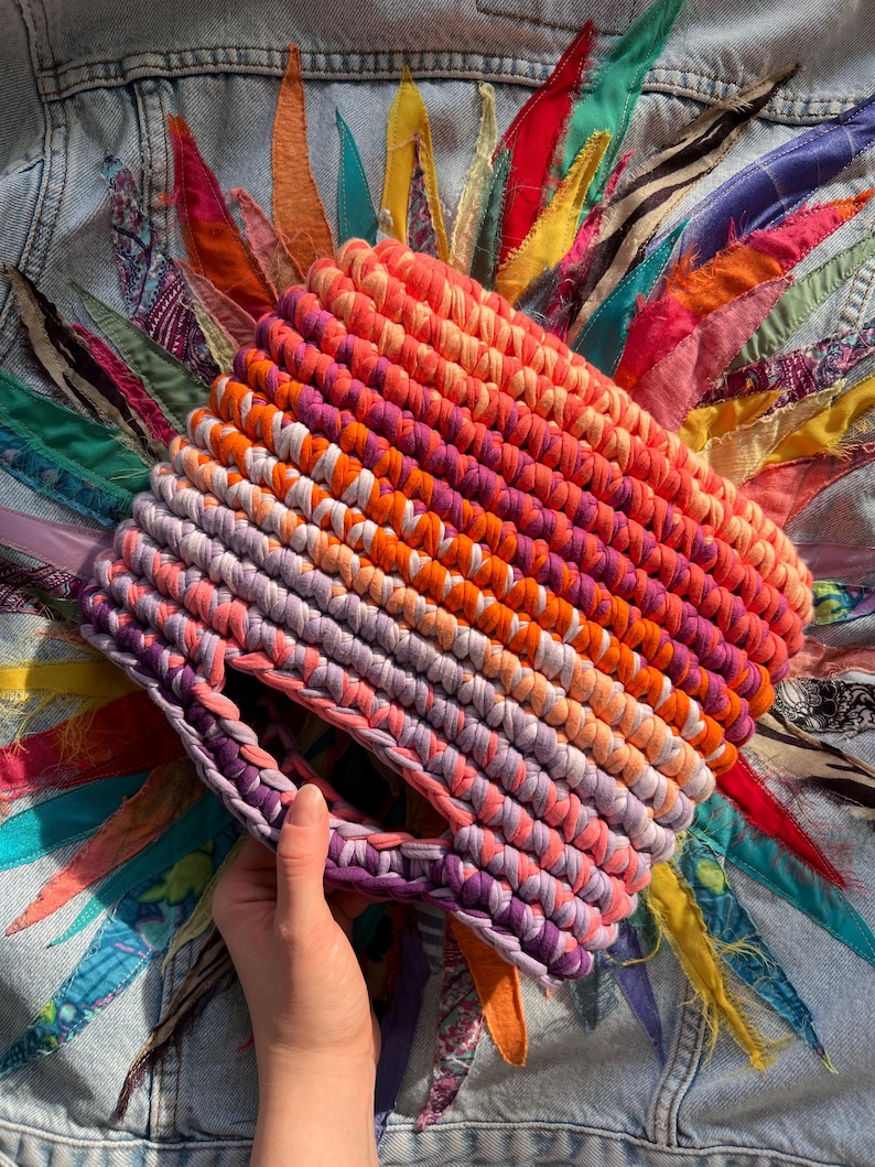 May include: A hand-crocheted bag made from thick fabric yarn featuring a vibrant gradient pattern that transitions from deep purple and lavender at the base to bright pink, orange, and coral tones at the top, with a circular integrated handle.