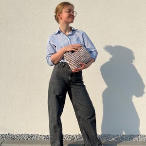 May include: A light blue button-down shirt with thin white stripes, dark gray denim jeans, and beige sneakers. A small, textured, light beige crocheted handbag with a thick, rounded handle is held in front of the torso.