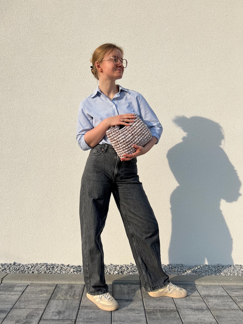 May include: A light blue button-down shirt with thin white stripes, dark gray denim jeans, and beige sneakers. A small, textured, light beige crocheted handbag with a thick, rounded handle is held in front of the torso.