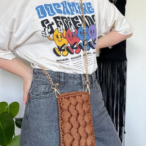 May include: A rectangular brown crochet phone pouch featuring a braided texture pattern. The pouch is attached to a gold-toned metal chain strap with lobster claw clasps, allowing it to be worn as a crossbody accessory for carrying a mobile device.