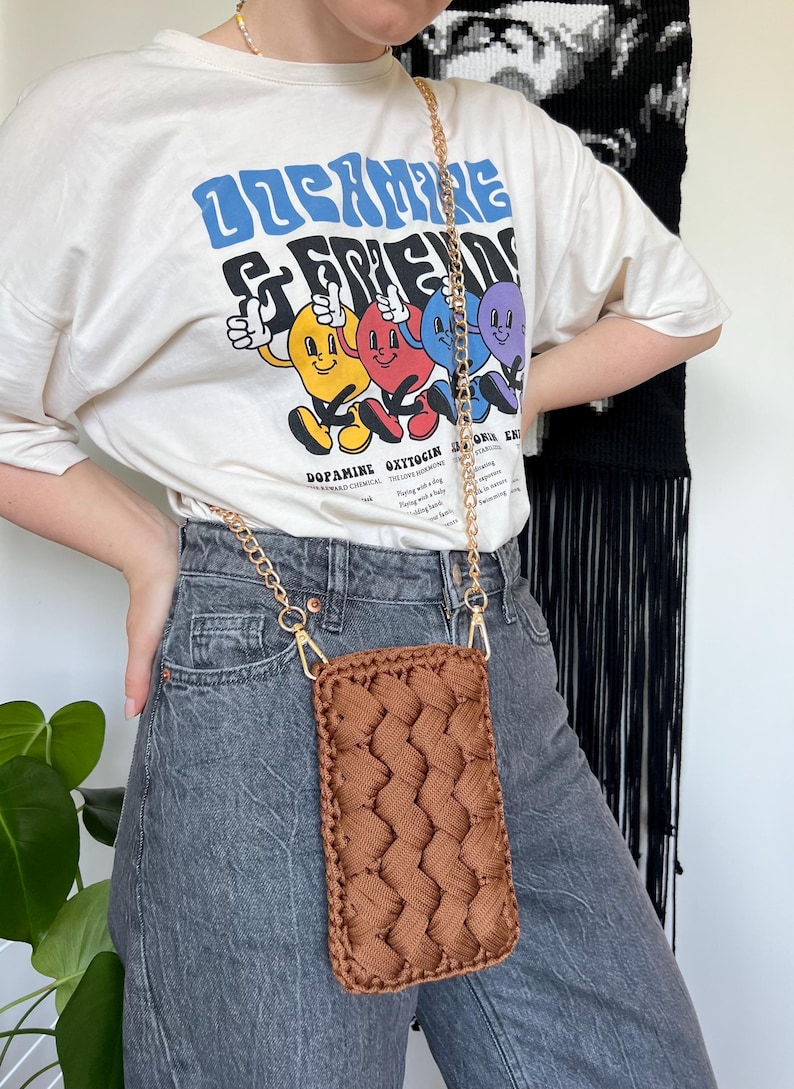 May include: A rectangular brown crochet phone pouch featuring a braided texture pattern. The pouch is attached to a gold-toned metal chain strap with lobster claw clasps, allowing it to be worn as a crossbody accessory for carrying a mobile device.
