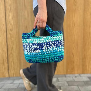 May include: A small, rectangular, hand-crocheted tote bag featuring a variegated color pattern of deep navy blue, bright turquoise, and white yarn. The bag has two integrated, thick crocheted handles and a dense, textured stitch construction.