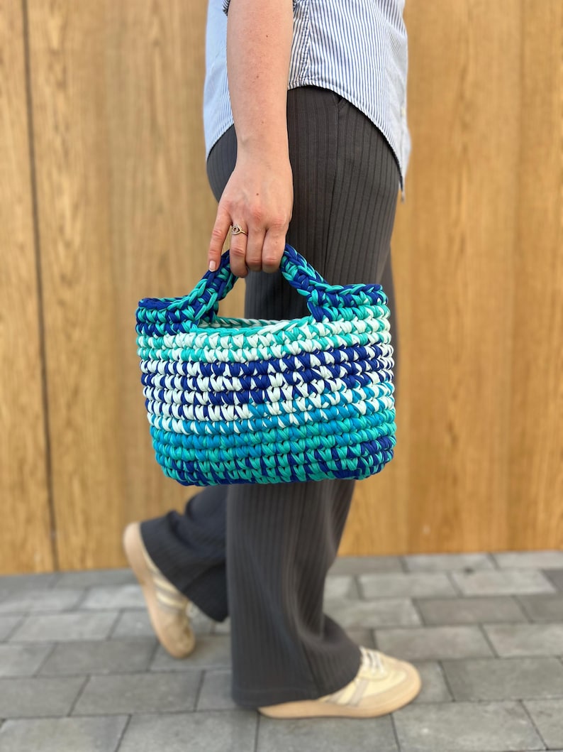 May include: A small, rectangular, hand-crocheted tote bag featuring a variegated color pattern of deep navy blue, bright turquoise, and white yarn. The bag has two integrated, thick crocheted handles and a dense, textured stitch construction.