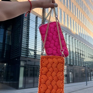 May include: Two rectangular crossbody bags featuring a thick, woven cord texture. One bag is vibrant hot pink, and the other is bright orange. Both bags are suspended by metallic gold-tone chain straps with lobster claw clasps at the top.