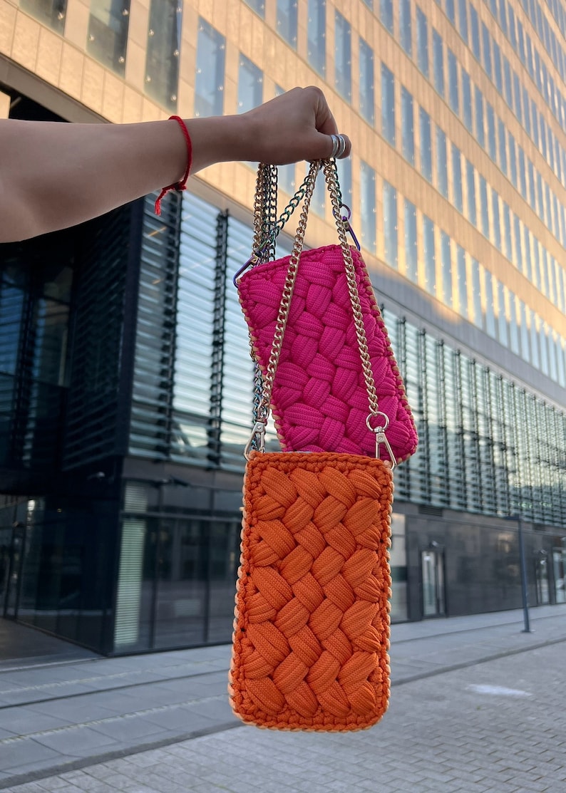 May include: Two rectangular crossbody bags featuring a thick, woven cord texture. One bag is vibrant hot pink, and the other is bright orange. Both bags are suspended by metallic gold-tone chain straps with lobster claw clasps at the top.