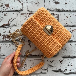 K&ouml;nnte beinhalten: Eine pfirsichfarbene H&auml;keltasche mit einem strukturierten, geknoteten Maschenmuster. Die Tasche verf&uuml;gt &uuml;ber einen goldfarbenen Metalldruckknopfverschluss und einen passenden goldfarbenen Kettenriemen mit einem dicken, geh&auml;kelten Schulterpolster.