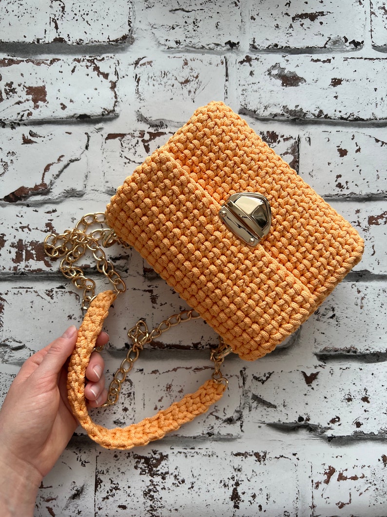 K&ouml;nnte beinhalten: Eine pfirsichfarbene H&auml;keltasche mit einem strukturierten, geknoteten Maschenmuster. Die Tasche verf&uuml;gt &uuml;ber einen goldfarbenen Metalldruckknopfverschluss und einen passenden goldfarbenen Kettenriemen mit einem dicken, geh&auml;kelten Schulterpolster.