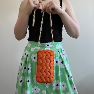 May include: A rectangular orange crochet crossbody bag featuring a thick braided texture and a gold-tone metal chain strap. The bag is constructed from cord-like yarn and has a structured, vertical orientation with a simple, clean design.