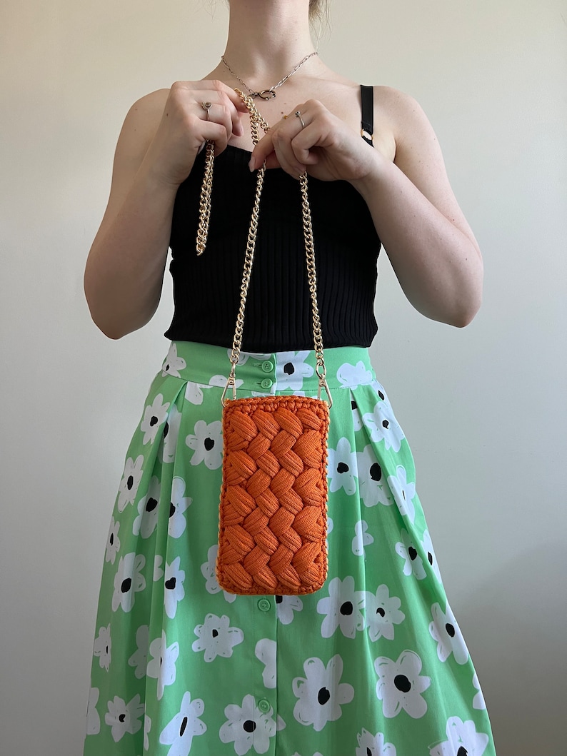 May include: A rectangular orange crochet crossbody bag featuring a thick braided texture and a gold-tone metal chain strap. The bag is constructed from cord-like yarn and has a structured, vertical orientation with a simple, clean design.