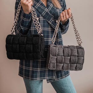 May include: Two rectangular handbags featuring a thick, woven knit texture. One bag is solid black and the other is dark gray. Both handbags include a chunky, metallic silver chain strap attached to the top, providing a sturdy handle for carrying.