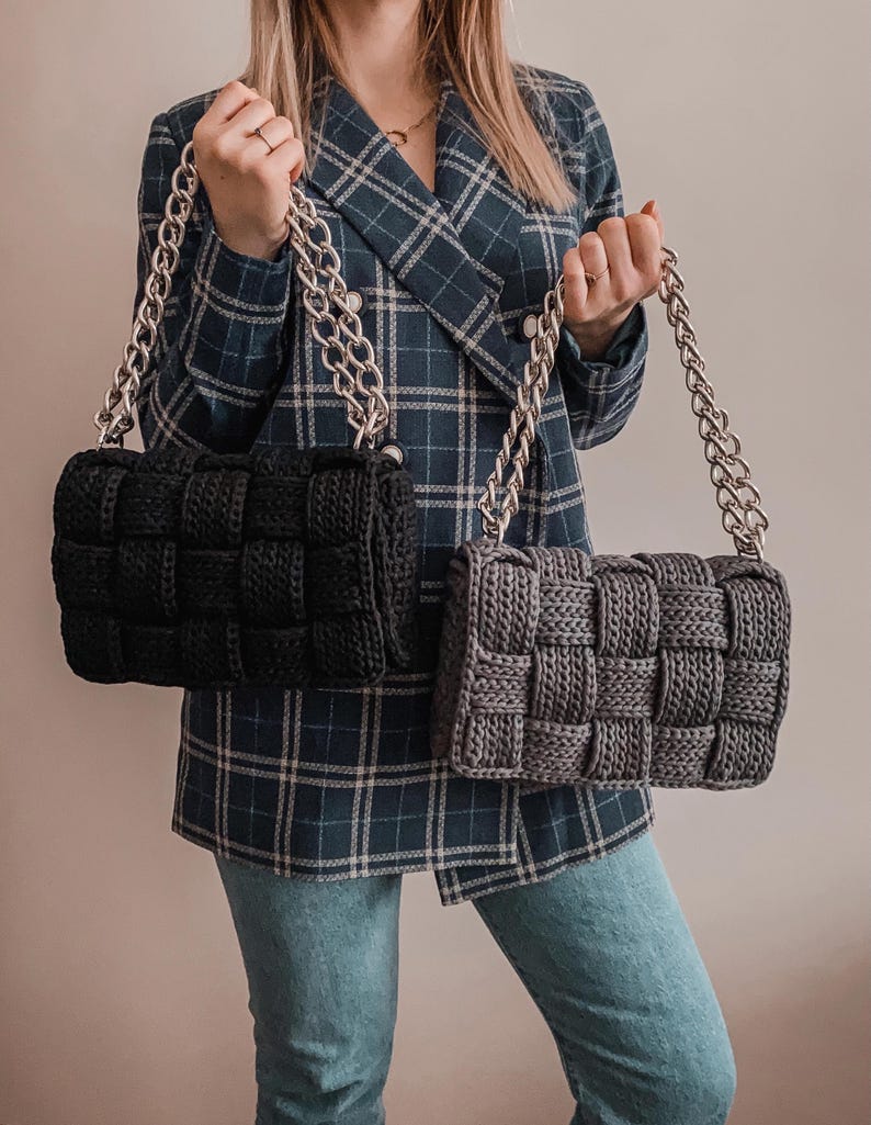 May include: Two rectangular handbags featuring a thick, woven knit texture. One bag is solid black and the other is dark gray. Both handbags include a chunky, metallic silver chain strap attached to the top, providing a sturdy handle for carrying.