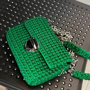 K&ouml;nnte beinhalten: Eine smaragdgr&uuml;ne H&auml;keltasche mit strukturierter rechteckiger Form und gewebter Textur. Die Tasche verf&uuml;gt &uuml;ber einen polierten silberfarbenen Metalldruckverschluss an der Frontklappe sowie einen passenden gr&uuml;nen H&auml;kelriemen mit silbernen Kettengliedern.