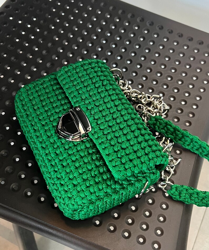 K&ouml;nnte beinhalten: Eine smaragdgr&uuml;ne H&auml;keltasche mit strukturierter rechteckiger Form und gewebter Textur. Die Tasche verf&uuml;gt &uuml;ber einen polierten silberfarbenen Metalldruckverschluss an der Frontklappe sowie einen passenden gr&uuml;nen H&auml;kelriemen mit silbernen Kettengliedern.