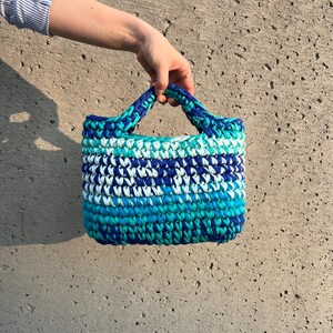 May include: A small, rectangular handbag crafted from thick, braided yarn in a variegated pattern of royal blue, turquoise, light blue, and white. The bag features two integrated, braided handles and a dense, textured crochet stitch construction.