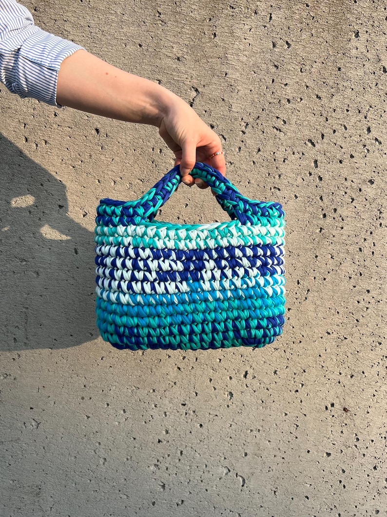 May include: A small, rectangular handbag crafted from thick, braided yarn in a variegated pattern of royal blue, turquoise, light blue, and white. The bag features two integrated, braided handles and a dense, textured crochet stitch construction.