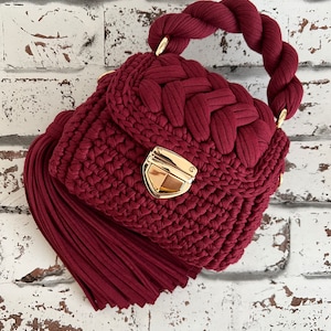 Puede incluir: Un bolso de ganchillo en color burdeos intenso con un asa trenzada gruesa, una borla grande a juego en el lateral y un cierre de presi&oacute;n de metal dorado pulido. El bolso est&aacute; hecho de cord&oacute;n de tela grueso con un patr&oacute;n de tejido texturizado.