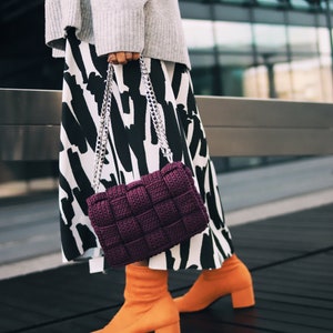 May include: A deep purple woven handbag with a silver chain strap, paired with a gray sweater, a black and white abstract print skirt, and bright orange block-heeled ankle boots. The handbag features a thick, square-patterned knit texture.