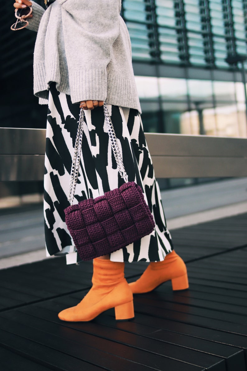May include: A deep purple woven handbag with a silver chain strap, paired with a gray sweater, a black and white abstract print skirt, and bright orange block-heeled ankle boots. The handbag features a thick, square-patterned knit texture.