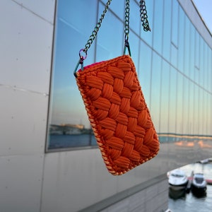 May include: A rectangular pouch crafted from thick, woven orange cord with a textured, braided pattern. The pouch features a light-colored stitched border and is suspended by a metallic, iridescent chain strap attached with matching hardware.